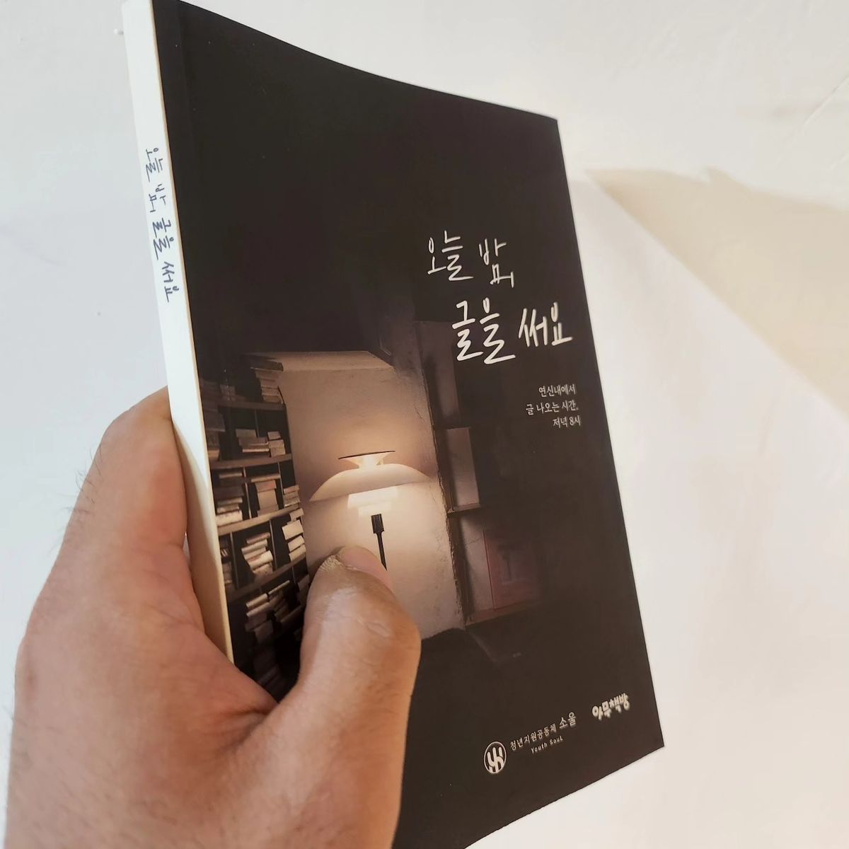 첫 책 「오늘 밤, 글을 써요」를 손에 든 모습. 검은 표지에 스탠드 불빛과 책장이 담겨 있어요.