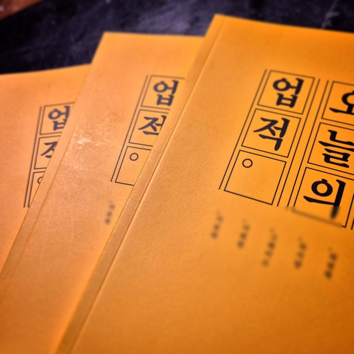노란 표지의 〈오늘의 업적〉 여러 권이 겹쳐 놓여 있어요.